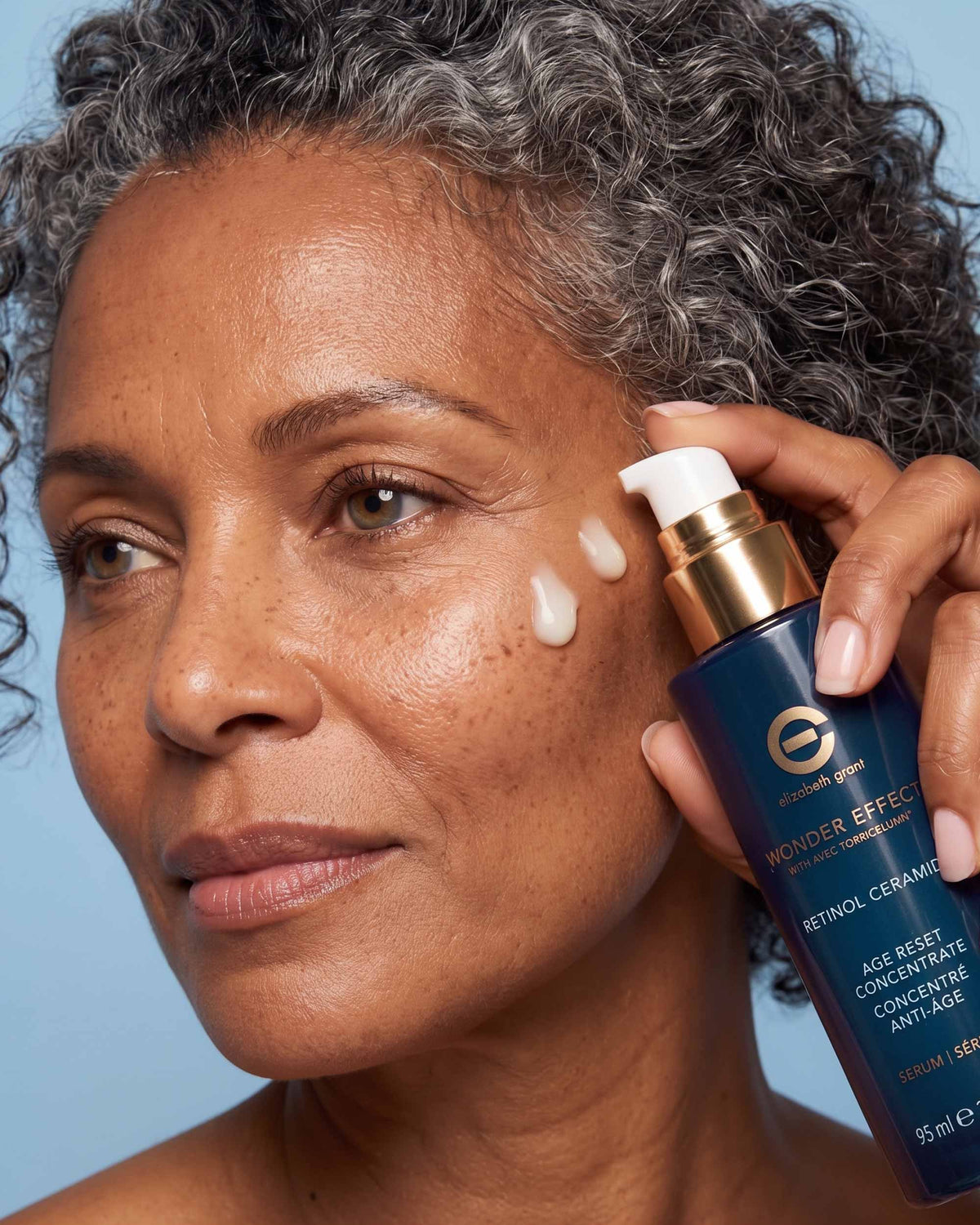 A close-up of a woman with gray curly hair applying cream from a blue and gold skincare pump bottle to her cheek against a light blue background.